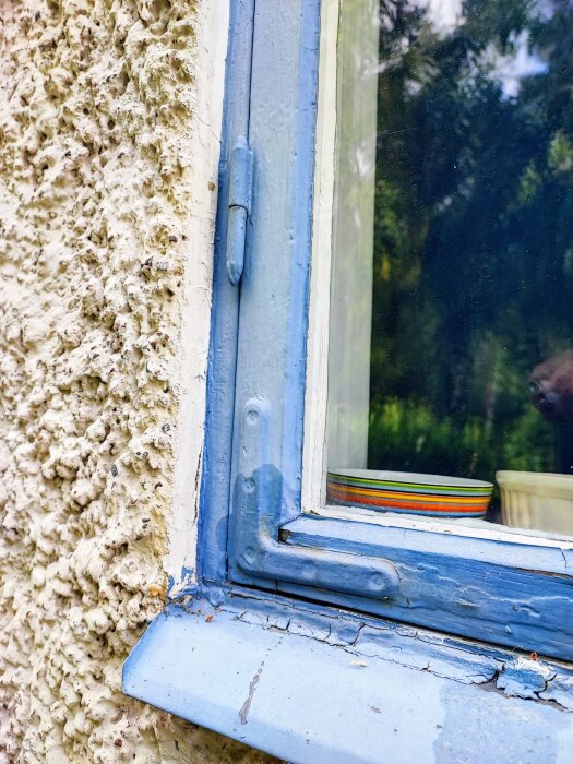 Närbild på ett gammalt fönster med blå karm och reveterad fasad; synliga sprickor och skador på fönsterkarmen.