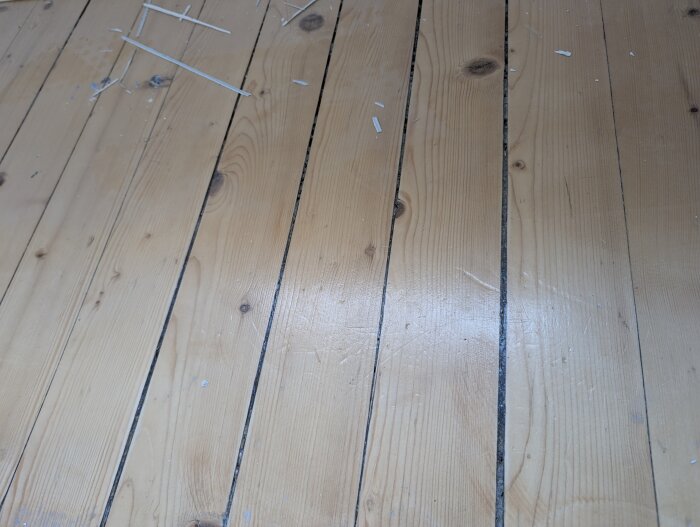Slitet obehandlat plankgolv med varierande mellanrum mellan brädor och några spån på ytan.