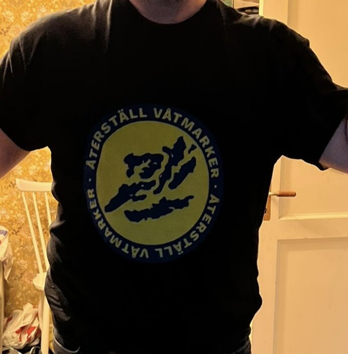 Person i svart t-shirt med en cirkellogga som har texten "Återställ våtmarker" runt ett blått motiv på gul bakgrund.