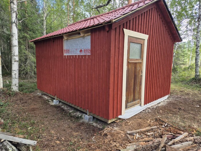Rött uthus 5x4 meter med träpanel och rött tak, beläget i skogsområde. Uthuset byggt med plastbandade spikar och Hitachi spikpistol.