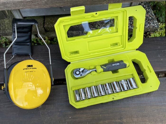 Gula Peltor-hörselskydd och en Ryobi spärrnyckelsats i en gul plastväska ligger på en träbänk utomhus.