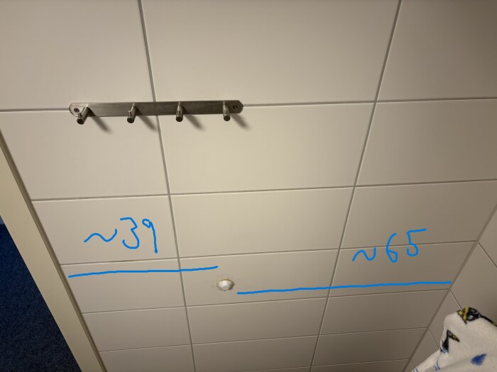 Vägg med kakel, kroklist och markerade avstånd i blått (~39 cm och ~65 cm) mellan vägg och elektrisk anslutning för handdukstork.