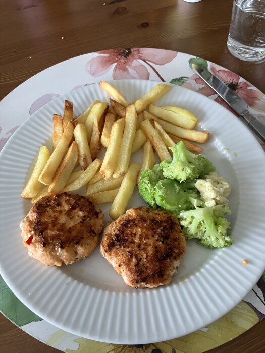 Tallrik med laxburgare, pommes frites och broccoli på blommig bordstablett.
