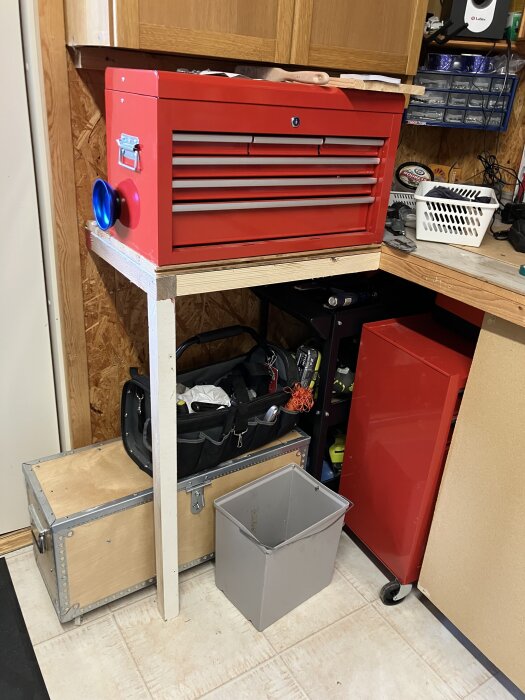 Röd verktygslåda på arbetsbänk med förvaring, verktygstavla och lådor, över ett trägolv bredvid låda och papperskorg.