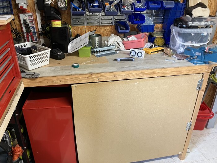 Arbetsbänk med verktygstavla och förvaringslådor, spånskiva fastsatt med gångjärn som dörr. Verktyg, tejp och förvaringslådor i bakgrunden.