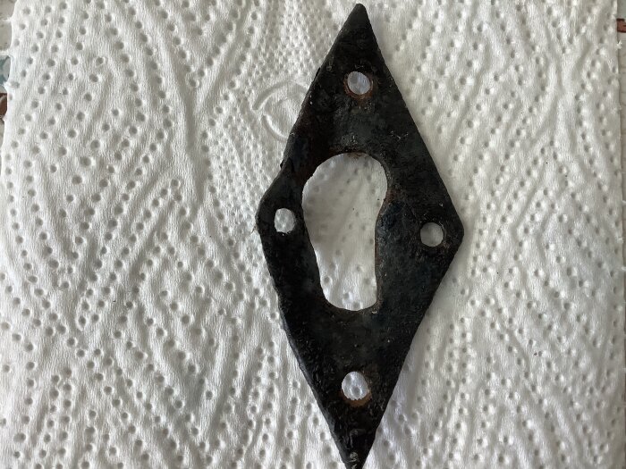 Rostig, diamantformad nyckelskylt med centralt nyckelhål vilar på vit servett. Fyra skruvhål syns runt kanten.