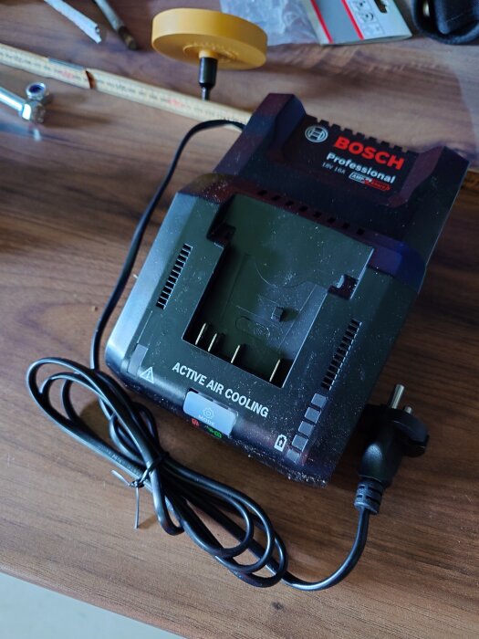 Bilden visar en Bosch batteriladdare med kabel och texten "Active Air Cooling" på ett träbord, omgiven av verktyg och tillbehör.