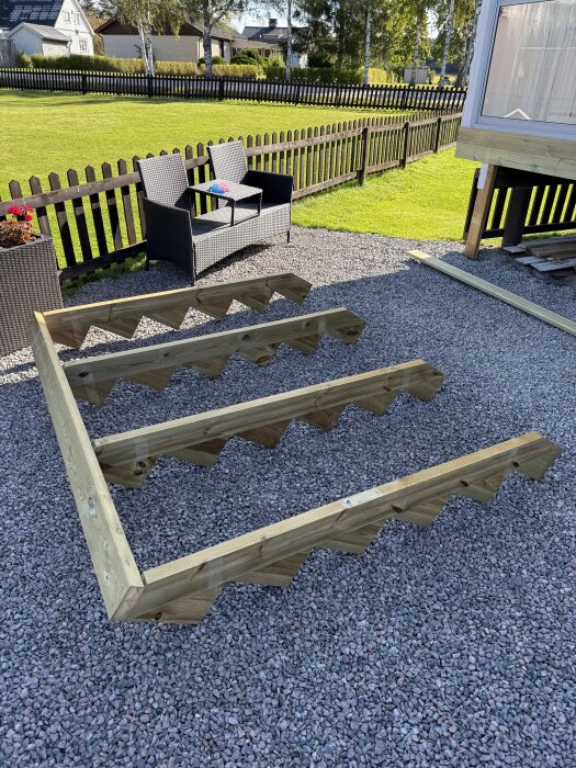 Trästomme för entrétrappa under byggnation, placerad på grus vid en husvägg, med trädgårdsmöbler och staket i bakgrunden.