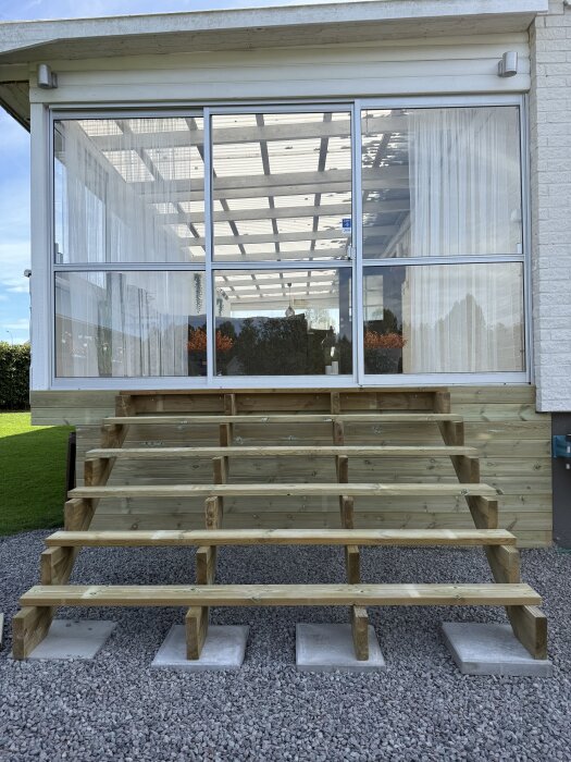 Bred entrétrappstomme i trä under konstruktion, placerad på stenlagd mark framför ett glasparti på ett hus.