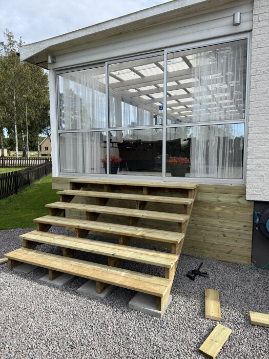 Bred trätrappa vid ingången till huset, med en stomme av trallbrädor, omgiven av grus och grönska, redo för installation av decklights.