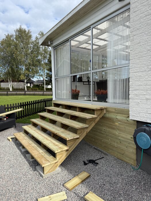 Bred trätrappa under konstruktion framför en inglasad veranda, trappstegen är ofärdiga och omgivna av verktyg och trallbrädor.