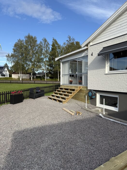 Bred entrétrappa under konstruktion vid ett vitt hus, med grusgång och sittplats bredvid.