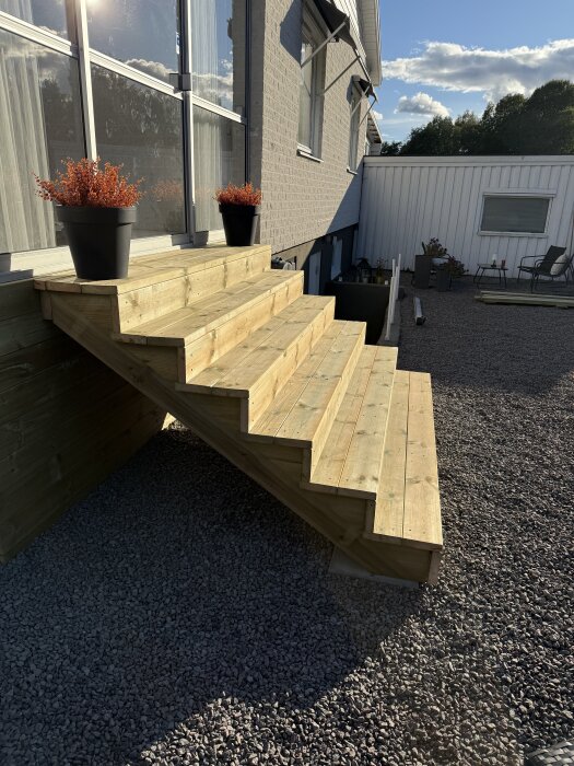 Nybyggd bredare entrétrappa av trä med svartmålade krukor vid sidorna, placerad mot ett hus med grusad mark runtom.