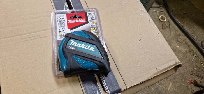 Makita 10m måttband i förpackning, placerat på en kartong i en verkstadsmiljö.