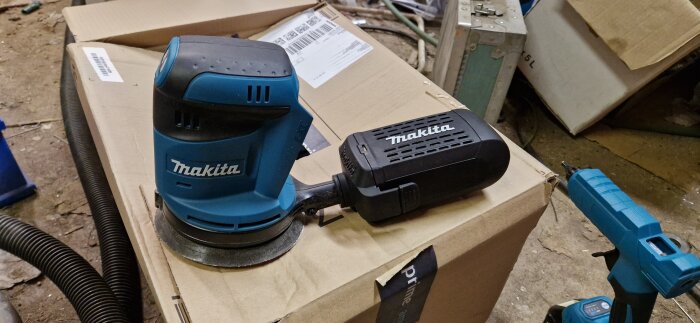 Makita slipmaskin och smältlimpistol på en kartong i en verkstadsmiljö.