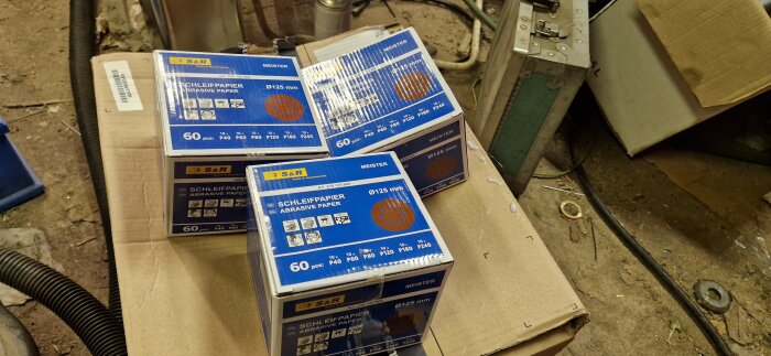 Förpackningar med S&R slippapper på en kartong i verkstadsmiljö.