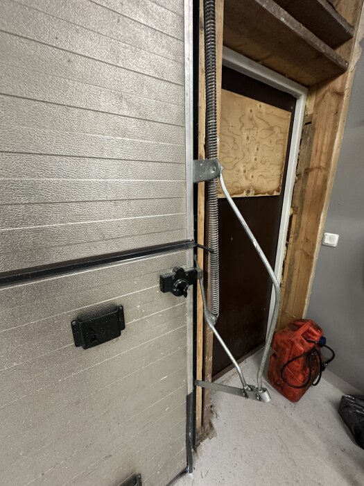 Gammal garageport i metall med fjädrar och mekanism synlig, intill en träpanel och en täckt dörr, verktyg och en orange plastbehållare.