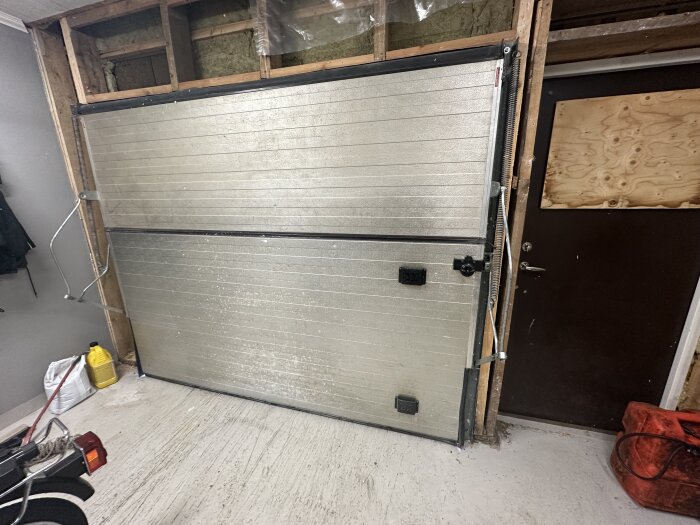 Gammal garageport i metall, delvis nedmonterad, med synlig isolering och verktyg runtomkring inne i ett garage.