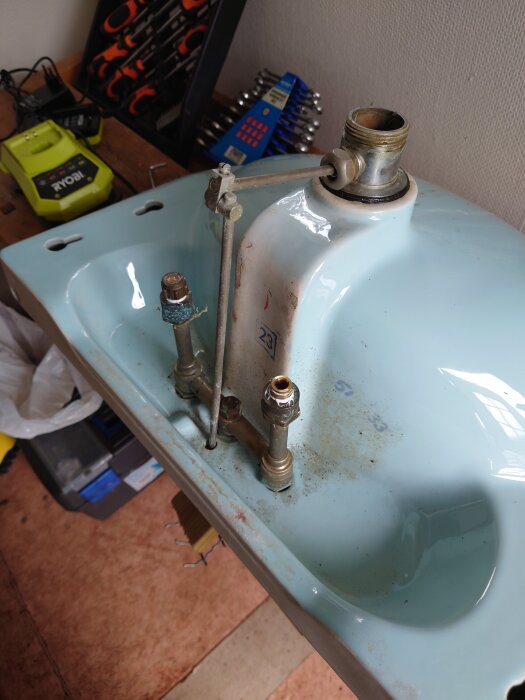 Ett handfat sett underifrån med synliga röranslutningar och verktyg i bakgrunden, kanske för reparation eller ommontering.