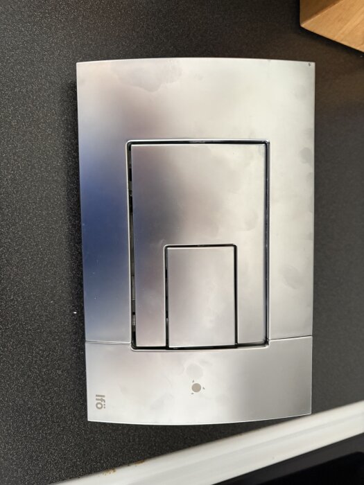 Kromfärgad dubbelspolknapp till Ifö Sign toalett, monterad på en svart yta.