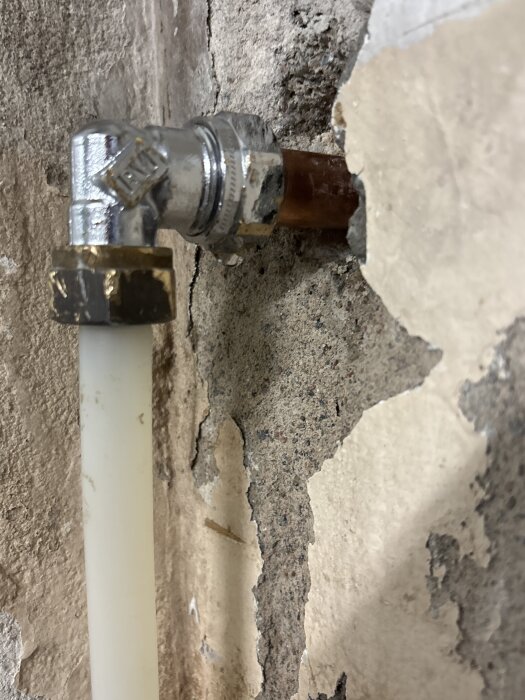Kopparrör med vit slang ansluten till vinklad metallkoppling mot sliten vägg, mindre läckage synligt.