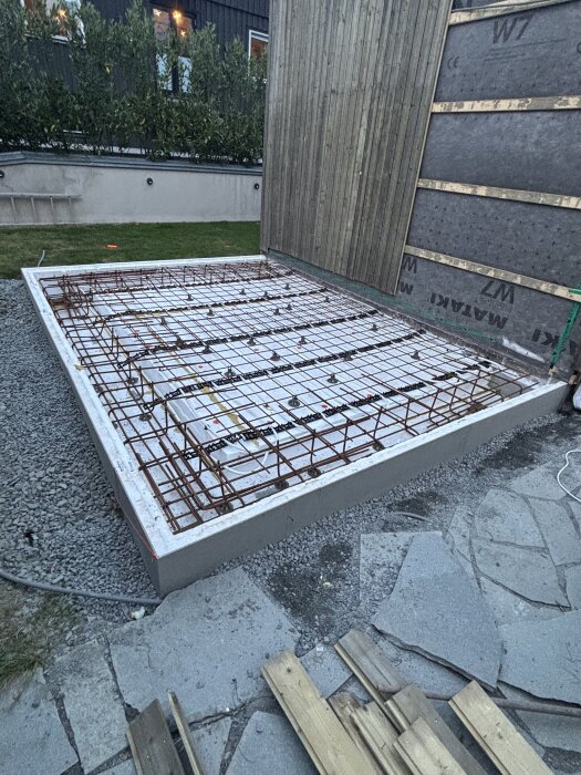 Armeringsjärn förberett på ett gjutform vid en byggnad, omgivet av grus och stenplattor, redo för betonggjutning.