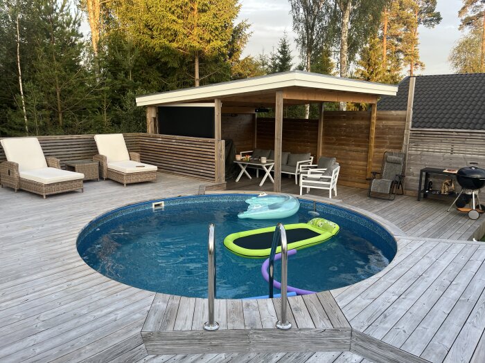 Poolområde med trädeck, solstolar, och ett enkelt tak. Poolen har leksaker i vattnet och en grill samt trädgårdsmöbler syns i bakgrunden.