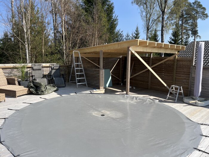 Uteplats med pågående bygge av tak och vägg, stegar, solstolar och presenning synliga under en klar himmel.
