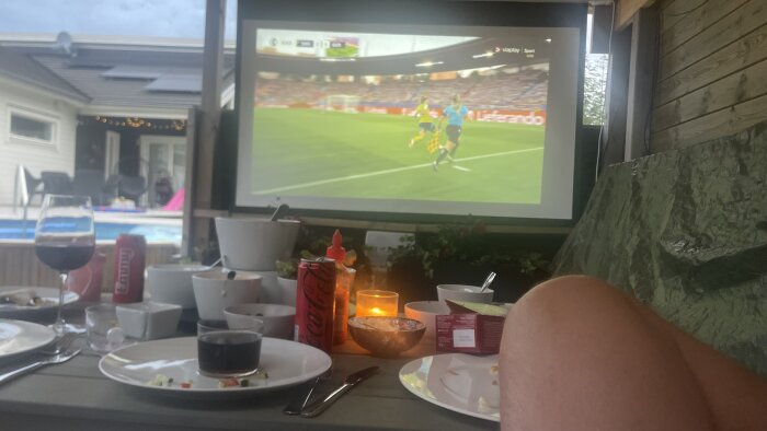 Utomhusprojektor visande fotbollsmatch, dukat bord med mat och dryck, pool och uteplats i bakgrunden.