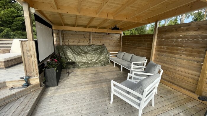 Takterrass med projektorduk, soffor och bord under trätak. Gröna växter i krukor och byggverktyg på golvet.