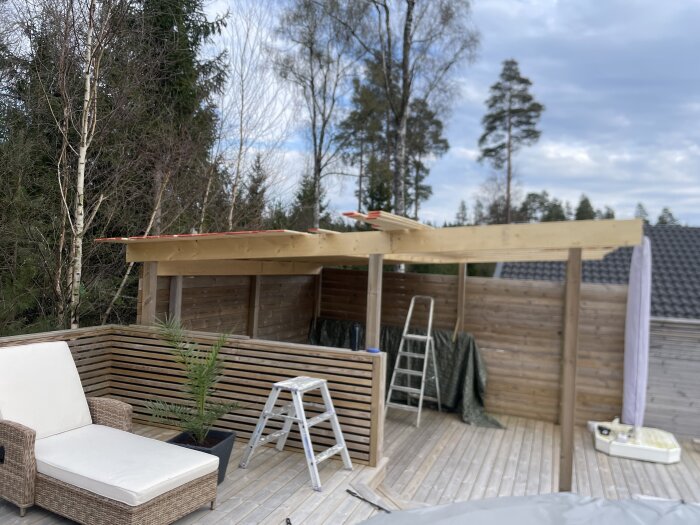 Byggprojekt pågår med en ny trästruktur på en altan, stege i förgrunden, omgiven av träd och modern utemöbel.