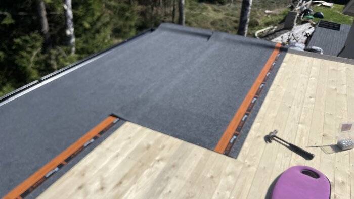 Takläggning med nylagt takpapp och träpanel samt verktyg som hammare och spikburk på byggarbetsplats.