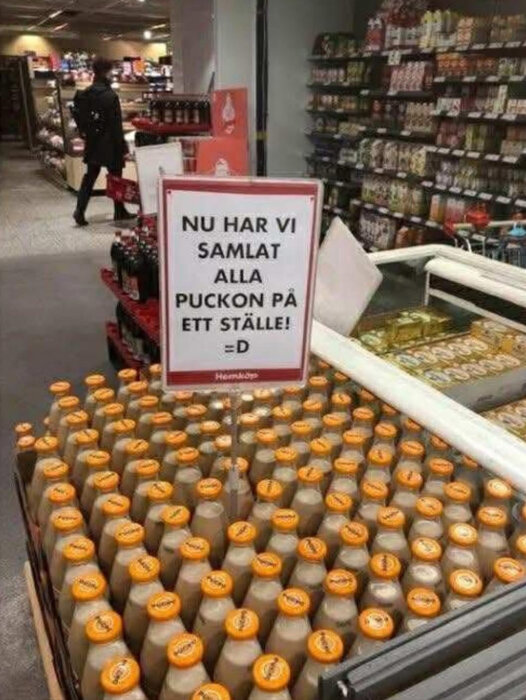 Skylt i en butik med texten "Nu har vi samlat alla puckon på ett ställe!" ovanför ett bord med läskflaskor med orange kapsyler.