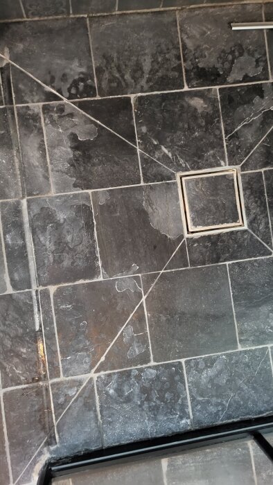 Fläckigt kalkstensgolv i dusch med mörka, matta plattor och vita märken runt golvbrunnen i ett badrum.