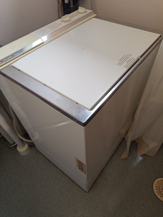 Äldre tvättmaskin med toppmatning i ett hörn av ett badrum, visas med panel och vred på ovansidan.