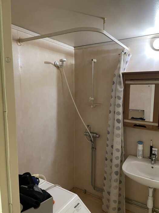 Badrum med duschhörna, duschdraperi med prickar, tvättmaskin och spegel. Rör målade med mattsvart lackfärg.