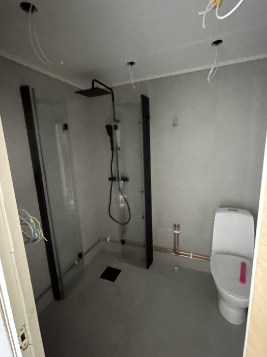 Modernt badrum med duschkabin, svartmålade rör och toalett. Taket har oslutna elinstallationer.