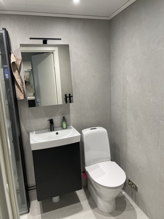 Modernt badrum med grå väggar, svartvitt handfat, spegel och toalett. Rör målade i mattsvart.