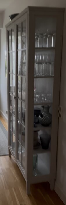 Vitrinskåp med glasdörrar och hyllor fyllda med glas och porslin, står på trägolv bredvid en ljus vägg.