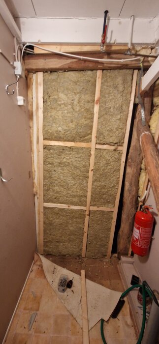 Vägg med glasfiberisolering i gammal lada, träreglar synliga, brandsläckare till höger, diverse verktyg på golvet.