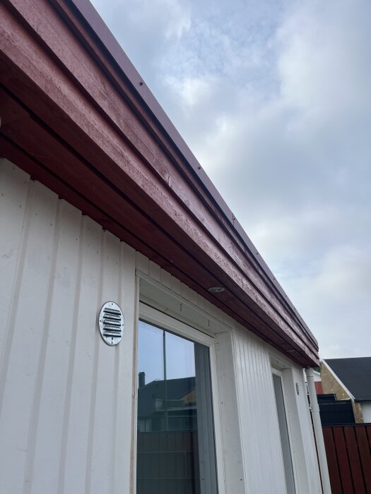 Fasad med avtagna fönsterfoder kring ett fönster på ett nybyggt hus, visande fria sidoväggar och ett rött fönsterbleck ovanför.