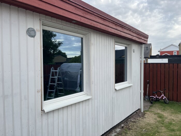 Vitt hus med två fönster utan foder. Fönsterblecken är monterade. En stege och en barncykel syns i bakgrunden.
