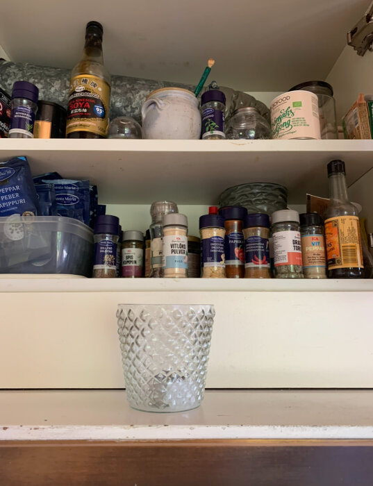 Kryddhylla i kök med olika kryddor och flaskor, samt en glasbehållare i förgrunden.