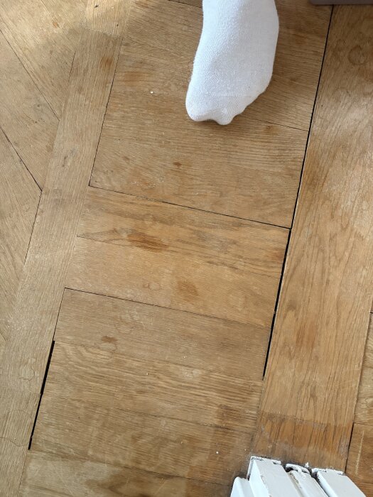 Fiskbensparkett i ek med sprickor och fläckar, vit socka synlig på golvet, våtmarkeringar och skavda ytor.