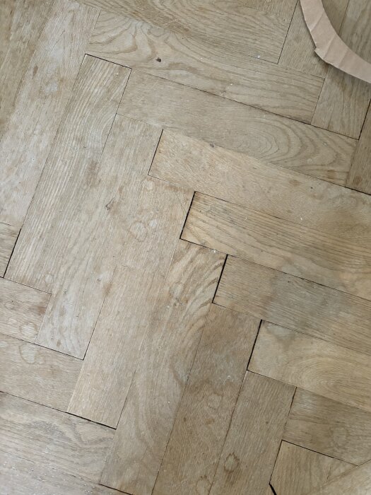 Fiskbensparkett i ek med märken och springor, gammal yta från lack eller fukt; synliga fettfläckar och slitage, matta toner och varierande nyans.