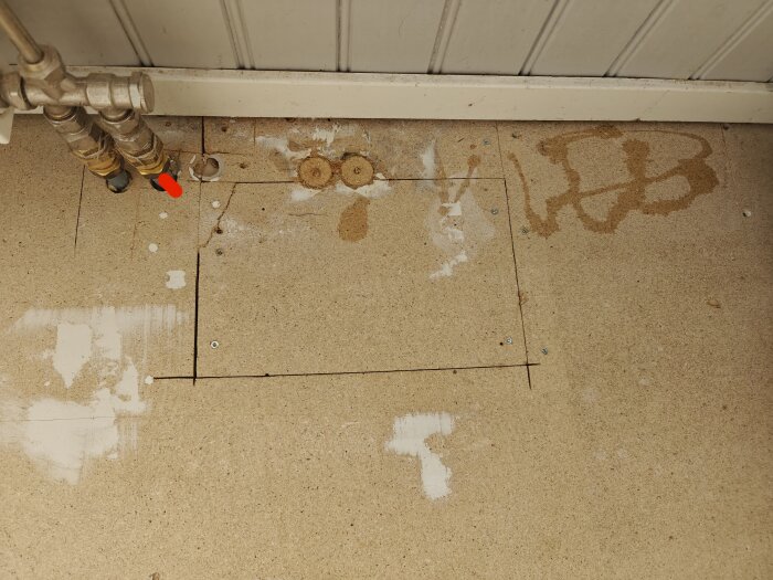 Fuktskadad spånskiva med markering för omsågning, synliga fläckar och skruvar under rör och närliggande vägglist i ett renoveringsprojekt.