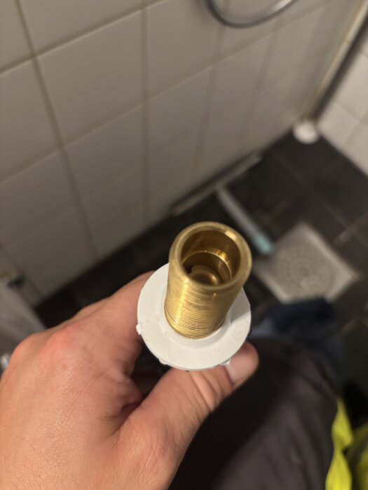 Hand som håller en mässingsanslutning med konisk kant och vit plastfläns, mot en bakgrund av kakel i badrumsmiljö.