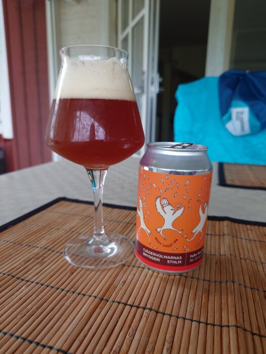 Ett glas skummande, brunaktig öl bredvid en färgglad ölburk på ett bord.