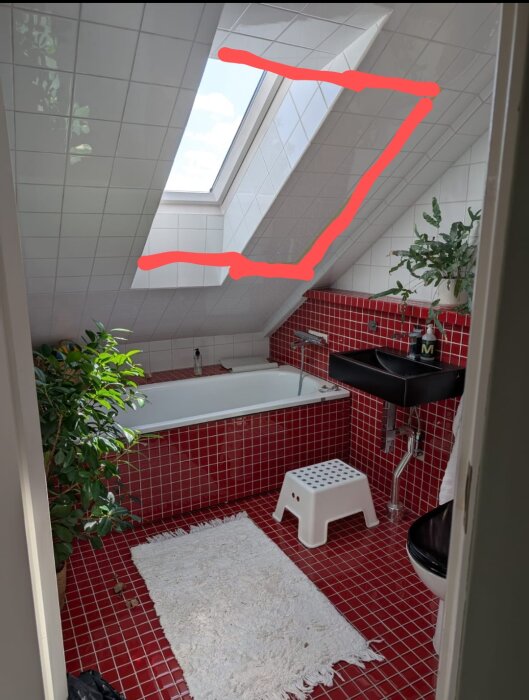 Trångt badrum med snedtak, rött kakel och inbyggt badkar. Markerat stort takfönster i vitt kakel, vit matta på golvet och en växt bredvid badkaret.