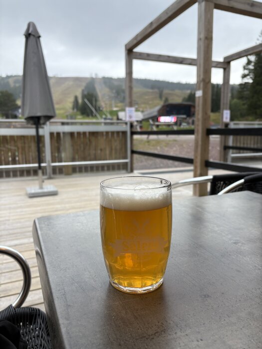 Ett glas öl med skum på toppen står på ett bord utomhus, omgivningen visar en uteservering med träd och ett berg i bakgrunden.